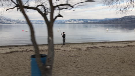 Aerial-View,-Young-Fisherman-With-Fishing-Rod,-Lakeside-Under-Snow-Capped-Hills-on-Cloudy-Winter-Day,-Orbit-Drone-Shot