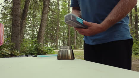 Man-spoons-coffee-grounds-into-base-of-Moka-Pot-on-table-at-campsite