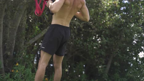 4K-Young-caucasian-fit-attractive-male-exersicing-outdoors-Trx-back-row-exercice-with-green-vegetation-background