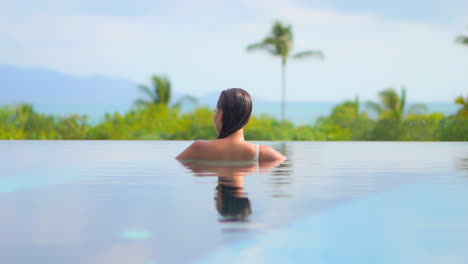 An-unrecognizable-woman-inside-infinity-pool-looking-at-amazing-tropical-greenery-on-hills-background-daytime,-body-reflection-in-water-surface