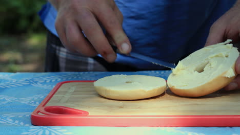 Close-up-of-man-making-bagel-sandwich-outdoors-on-picnic-table-while-camping