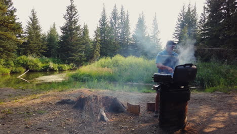 Wide-angle-view-of-man-grilling-steaks-while-camping-by-small-pond