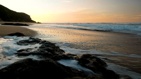Seawater-streaming-back-down-rock-on-beach,-golden-sunrise