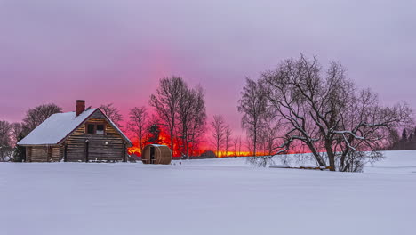 Timelapse-shot-of-cold-winter-snow-covered-day-in-rural-scenery-with-sunrise-in-the-background-over-wooden-cottage