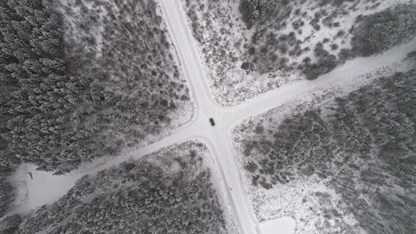 Aerial-top-down-of-car-on-snowy-rural-junction-surrounded-by-snow-covered-forest-trees-in-nature---orbiting-shot