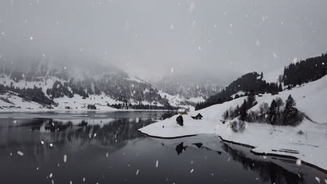 Snowy-foggy-mountain-landscape-reflects-in-clear-lake-water-during-snowfall,-aerial-drone-view
