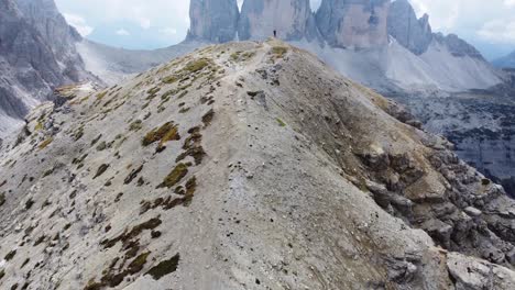 An-incredible-view-of-a-man-standing-alone-on-a-rock-formation-overlooking-Tre-Cime-di-Lavaredo-with-clouds-in-the-background