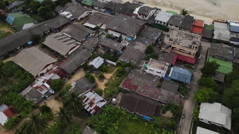 Drone-captures-the-aerial-view-of-many-homes-in-the-town-near-to-Malibu-Beach-on-Koh-Phangan,-Thailand,-along-with-a-boat-in-the-water,-a-sandy-beach,-corals,-and-mountains