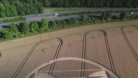 Cinematic-aerial-footage-of-crop-circles-in-a-field-of-yellow-grain-captured-by-drone-in-the-UK's-Micheldever-Station-with-moving-cars-nearby