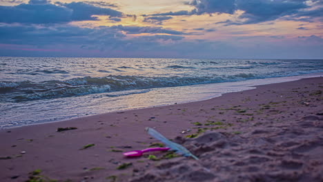 A-feather-and-a-child's-toy-sand-shovel-on-the-beach-with-a-cloudscape-above-the-waves-at-sunset---time-lapse