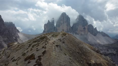 A-slow-moving-drone-shot-flying-away-from-a-hiker-standing-alone-in-the-mountains-of-the-Dolomites-in-South-Tyrol-in-Italy---He-is-looking-at-the-outstanding-rock-formations-of-Tre-Cime-di-Lavaredo
