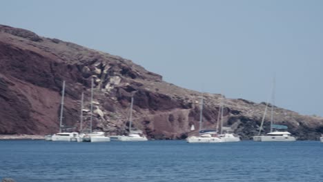Boats,-cliffside,-red-sand,-beach,-shore,-floating,-tourists,-Mediterranean,-ocean,-Santorini,-Greece