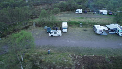 Aerial-dolly-shot-of-a-caravan-camp-site-with-motorhomes-and-vans-parked-in-late-evening-and-people-preparing-their-camp-site