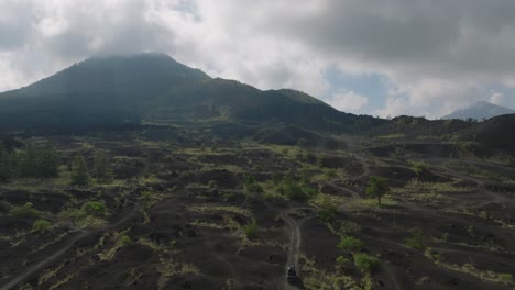 Black-lava-field-below-mount-Batur,-jeep-driving-on-sand-path,-aerial
