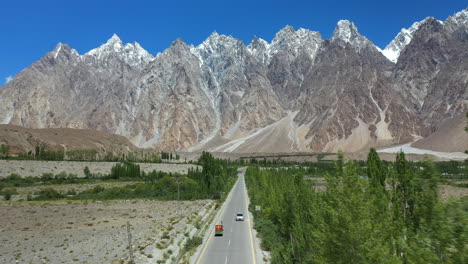Cinematic-drone-shot-of-Passu-Cones-in-Hunza-Pakistan,-moving-towards-the-Tupopdan-Peak-with-a-Tuk-Tuk-driving-on-Karakoram-Highway,-wide-aerial-shot