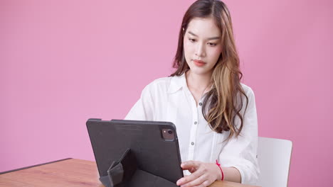 Junge-Asiatische-Frau,-Die-Mit-Einem-Digitalen-Tablet-Büro-Im-Studio-Mit-Rosafarbenem-Hintergrund-Spielt