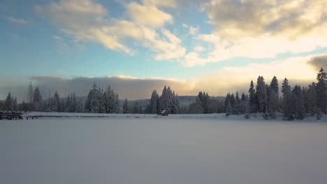 Retroceso-Aéreo,-Lago-Congelado-Cubierto-De-Nieve-Espesa-Durante-La-Temporada-De-Invierno-En-El-Bosque-De-Pinos-Durante-El-Amanecer,-Nubes-Dramáticas