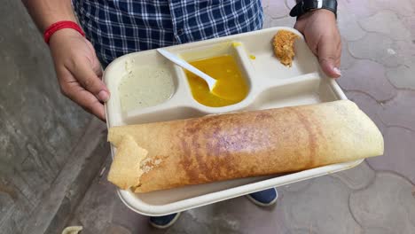 Masala-Dosa,-a-popular-South-Indian-dish-prepared-of-rice,-lentils,-potatoes,-Methi,-and-curry-leaves-and-served-with-chutneys-and-sambar,-is-being-held-in-a-man's-hand