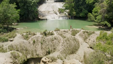 Aerial-is-tilting-up-showing-a-woman-walk-next-to-Roberto-Barrio-Waterfalls-in-Palenque,-Chiapas,-Mexico