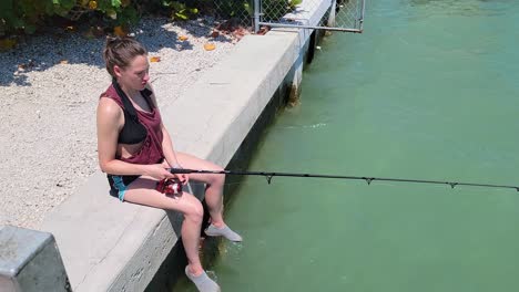 Woman-Fishing-while-Sitting-on-Edge-of-Pier-on-Shore-while-she-Reels-Spinning-Rod-and-Smiles-at-Camera