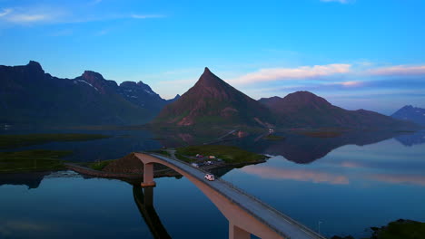 Aerial-following-shot-of-a-converted-van-driving-over-famous-bridges-Fredvang-on-Lofoten-Islands-with-Volandstind-reflecting-on-the-water