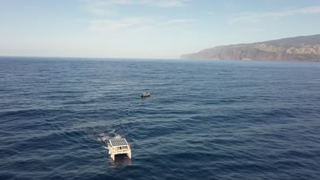 Drohnenaufnahme-Eines-Weißen-Katamarans-Mit-Sonnenkollektoren,-Der-Von-Einem-Kleineren-Boot-An-Der-Küste-Madeiras-Wegfährt