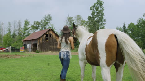 Attractive-young-cowgirl-on-farm-looks-off-into-the-distance,-then-walks-her-horse-back-to-the-barn