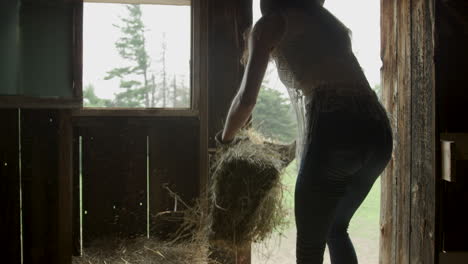 Stylish-young-cowgirl-in-a-barn-tosses-straw-down-to-her-horse