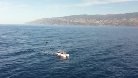 Drohnenaufnahme-Eines-Weißen-Katamarans-Mit-Sonnenkollektoren,-Der-Von-Einem-Kleineren-Boot-An-Der-Küste-Madeiras-Wegfährt-2