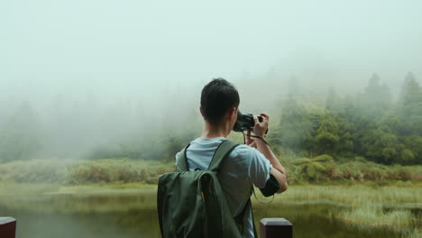 A-young-Asian-male-photographer-in-a-white-T-shirt-and-a-backpack-is-taking-photographs-of-nature-by-a-lake-in-the-mountains,-in-Taiwan