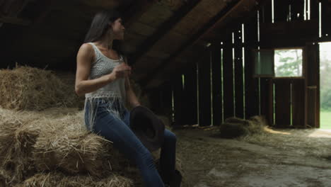 Confident-young-cowgirl-in-a-barn-puts-on-her-cowboy-hat-and-gloves,-picks-up-a-bale-of-straw-and-carries-it