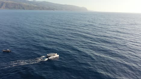 Drohnenaufnahme-Eines-Weißen-Katamarans-Mit-Sonnenkollektoren,-Der-Von-Einem-Kleineren-Boot-An-Der-Küste-Madeiras-Wegfährt-1