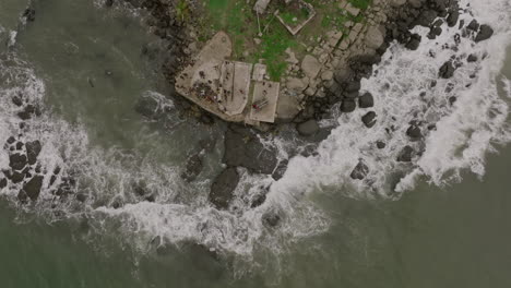 Slow-motion-aerial-footage-top-down-of-ocean-waves-crashing-into-rocks-with-group-of-people-at-the-point-of-a-peninsula