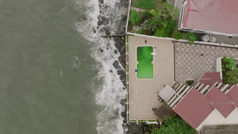 Aerial-top-down-footage-that-shows-a-pool-at-a-hotel-that-is-next-to-the-ocean,-with-waves-crashing-against-the-rocks-in-Sierra-Leone