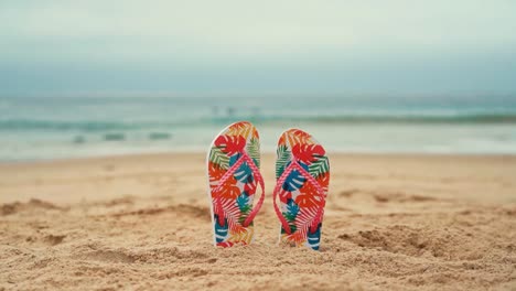 Colourful-flip-flops-buried-in-the-sand-with-the-sea-in-the-back,-Beach-vacations-and-travel-background-with-no-people