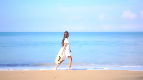 Attractive-Woman-in-White-Dress-With-Hat-in-Hand-Walking-on-Sandy-Beach-in-Front-of-Tropical-Sea,-Slow-Motion