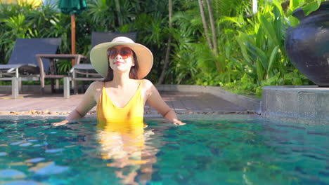 A-pretty-young-woman-in-a-yellow-bathing-suit-sunglasses-and-a-sun-hat-leans-along-the-back-edge-of-a-resort-swimming-pool