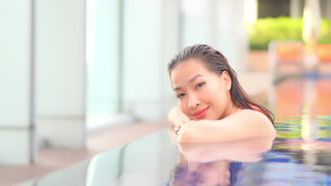 A-Close-up-of-an-attractive-young-woman-leaning-her-chin-on-her-folded-arms-along-the-edge-of-an-infinity-edge-pool-turns-her-head-and-smiles-at-the-camera