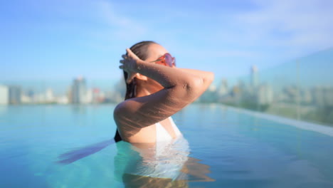 With-a-modern-skyline-behind-her-a-close-up-of-a-young-woman-in-a-rooftop-swimming-pool-smoothing-back-her-wet-hair