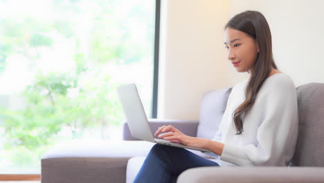 Close-up-of-a-young-woman-sitting-on-a-couch-typing-on-the-laptop-balanced-on-her-legs