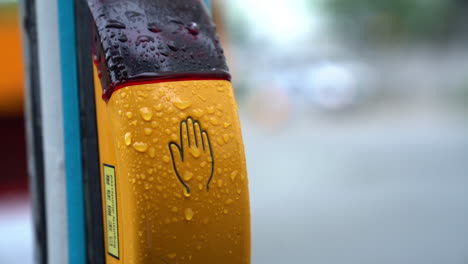 Button-for-traffic-light-at-the-crossroads-with-blurred-car-in-background-during-a-raining-da