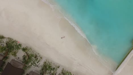 Una-Niña-Tendida-En-La-Orilla-De-La-Playa-De-Una-Isla-De-Maldivas,-Agua-Azul-Y-Vegetación-Verde