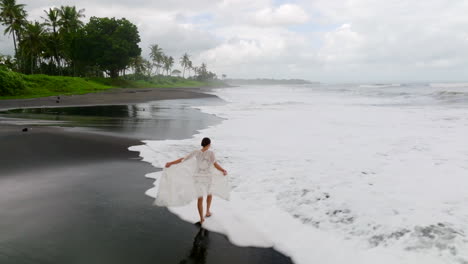 Low-aerial-shot-of-young-woman-in-bikini-walking-alone-along-a-black-sand-beach-shoreline