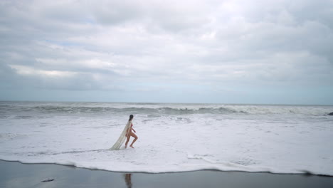 Woman-in-bikini-and-long-white-cover-walks-slowly-along-a-moody,-cloudy-beach