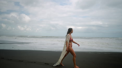 Attractive,-young,-independent-woman-in-bikini-walks-on-black-sand-beach-alone-in-Bali,-Indonesia