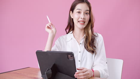 Happy-exuberant-Asian-woman-in-white-clothing-sitting-on-a-white-chair-talking-slowly-while-using-an-electronic-tablet-against-an-isolated-pink-background