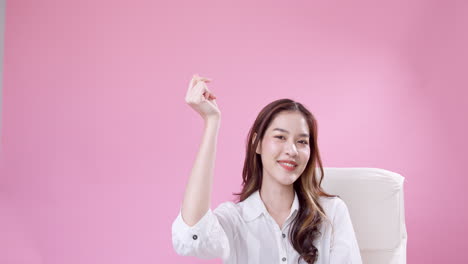 Happy,-delighted-Asian-woman-sitting-on-a-white-chair-and-snapping-her-finger-in-the-air-against-a-pink-background
