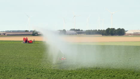 Landwirtschaftliches-Bewässerungssystem,-Das-Wasser-über-Pflanzen-Sprüht,-Windturbinen-Im-Hintergrund,-Zeitlupe-2