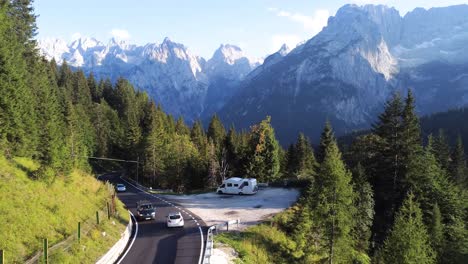 A-drone-shot-of-a-mountain-road-in-the-Dolomites,-with-cars-passing-by-and-an-auto-camper-parked-at-the-side-of-the-road-in-the-middle-of-a-pine-tree-forest-surrounded-by-mountains-in-north-Italy