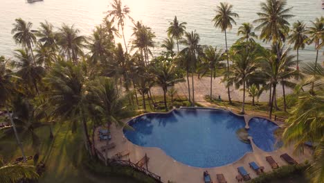 A-tropical-4K-drone-shot,-flying-in-between-palm-trees-with-a-view-of-a-charming-beach-resort-and,-the-ocean-and-a-swimming-pool,-at-sunset-hours-on-the-island-of-Koh-Kood-in-Thailand-in-SE-Asia